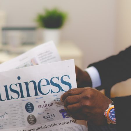 A person in a business suit is reading a newspaper with the headline "business," focusing on financial news and market trends. Only the person's hands and the newspaper are visible, symbolising the art of negotiating.