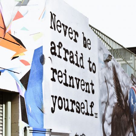 A colourful mural on a building-inspiring phrase wall with the text "Never be afraid to reinvent yourself" in bold, black letters shows your personal development. The mural includes abstract and figurative elements.