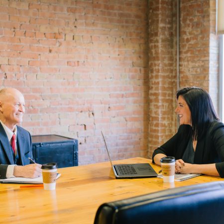 Two professionals, a man and a woman, are managing a meeting. They are conversing at a table with laptops and coffee cups in a modern office with brick walls.