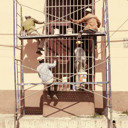 Three workers Working at Height on a scaffold painting and repairing the exterior wall of a building, under bright sunlight.
