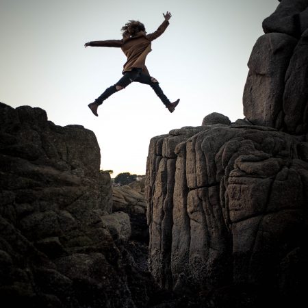 A person with curly hair, wearing a brown shirt and blue jeans, shows she is risk assessing before energetically jumping between two large, rugged rock formations under a soft evening sky. Symbolising Introduction to Risk Assessment.