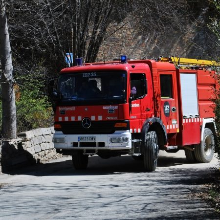 A red fire truck with the number "10235" and emergency lights on drives down a narrow road to be met by a fire marshal at a burning building.