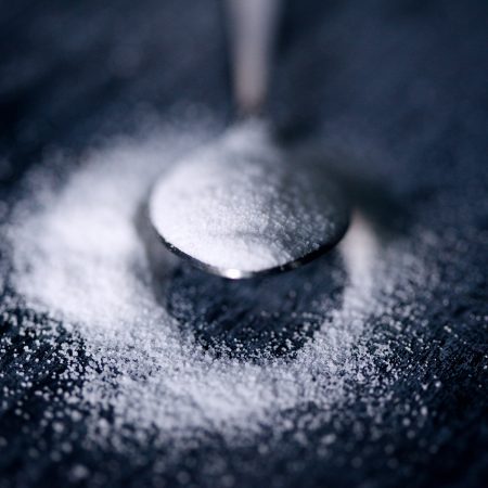 Close-up of a spoon sprinkling white sugar onto a dark surface, symbolising diabetes awareness, highlighting the granules' texture and the contrast between the sugar and the background,