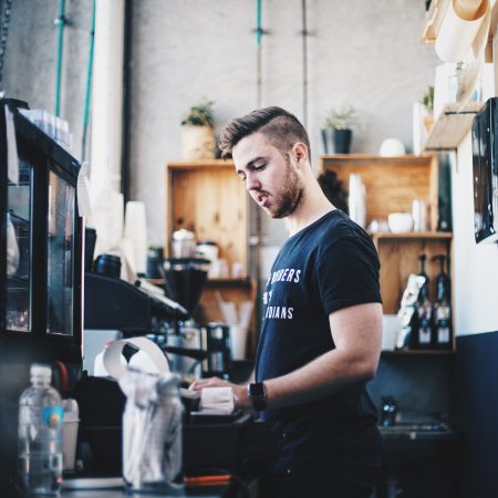 A man in a black t-shirt that says "brewers not baristas" stands inside a coffee shop, lone working near an espresso machine. The shop has a modern, industrial vibe with items lined up on shelves.