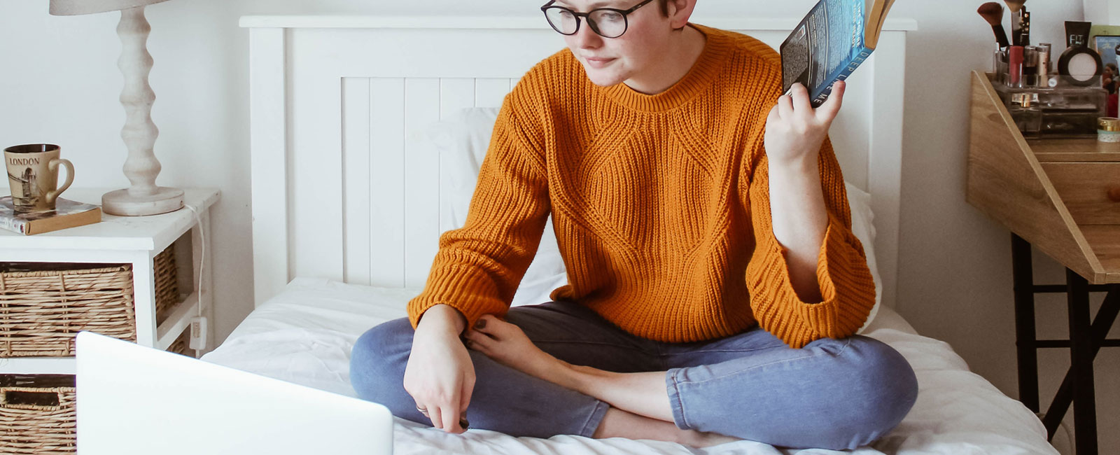 A person sitting cross-legged on a bed, wearing glasses and a burnt orange sweater, reading a book with a laptop beside them in a cozy bedroom setting.