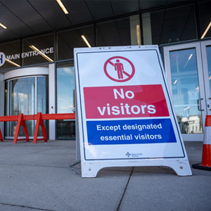 COVID-19 Safe Work Places should show a sign in front of a building entrance that reads "No visitors except designated essential visitors," featuring a graphic of a person with a prohibition symbol over it.